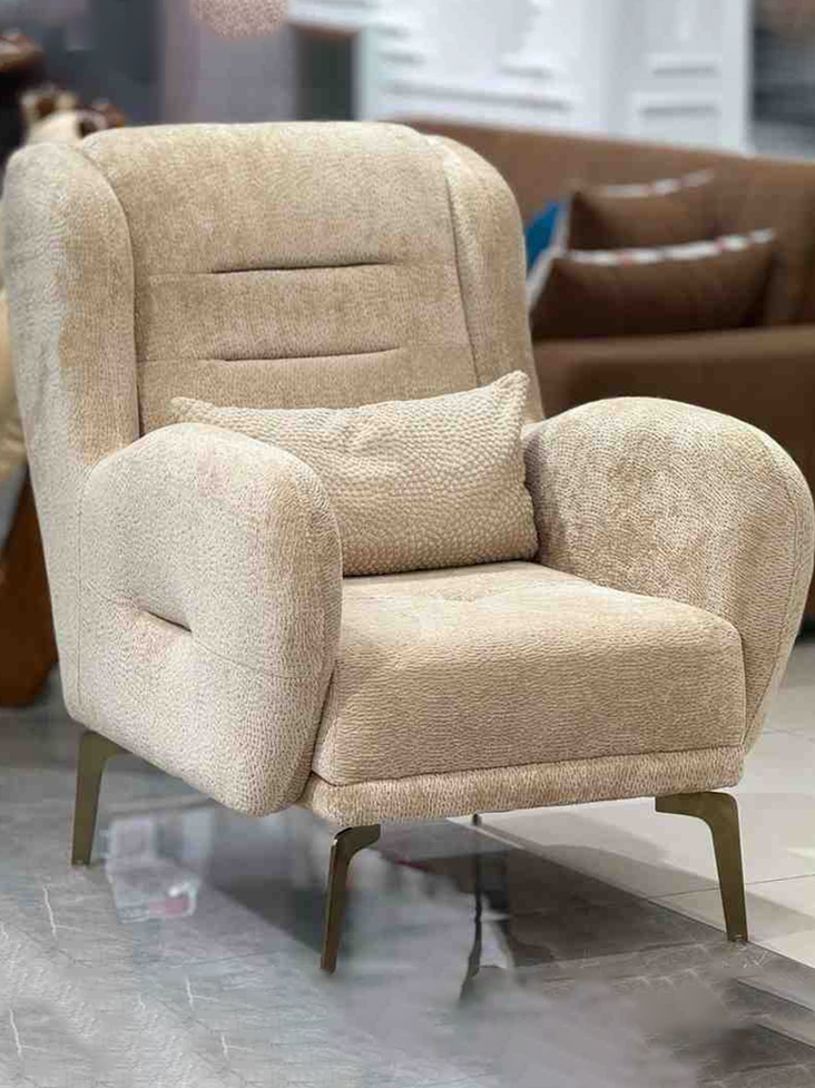 A beige upholstered armchair with a lumbar pillow and modern metallic gold legs.
